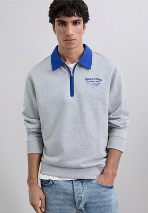 Joven vestido con un pulóver gris claro, cuello y cremallera azul, texto azul en el pecho y pantalones de mezclilla azul claros, de pie con una mano en el bolsillo.