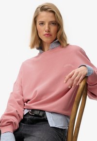 Różowy, oversize'owy sweter z prążkowanymi mankietami, nałożony na niebieską i białą koszulę w paski z kołnierzykiem. Model siedzi, trzymając drewniane krzesło.