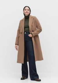 Ullblandad lång kappa i kamelfärg, dubbelknäppt design, kombinerad med en grön polotröja och utsvängda denimjeans.