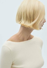 Coupe courte au carré avec une couleur blond clair, une texture lisse et une vague subtile. Portée avec un haut ajusté en maille côtelée de couleur blanc cassé et un fond neutre.