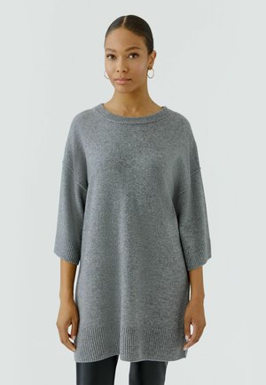 Femme aux cheveux attachés en chignon, portant des boucles d'oreilles créoles dorées et un pull en maille gris ample avec manches trois-quarts et détails côtelés.