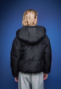 Musta, pöyheä huppari, jossa tikattu kuvio ja kiiltävä materiaali. Takana näkyvät saumat ja istuvuuden kontuurit.