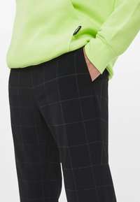 Person med svarta rutiga byxor, handen i fickan, och en limegrön långärmad skjorta med en synlig varumärkesetikett nära fållen.