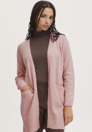 Cardigan rosa chiaro realizzato in morbido materiale peloso, con apertura frontale, maniche lunghe e tasche laterali. Indossato sopra un dolcevita marrone.