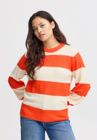 Pleteni pulover z širokimi oranžnimi in kremnimi črtami. Rebrast izrez in manšete. Mehak material, sproščen kroj. Spodaj nosite modre jeans.
