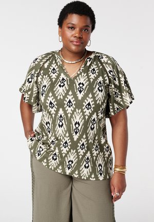 Femme aux cheveux courts et bouclés portant un chemisier vert et blanc à motifs, un pantalon olive, des bijoux en or, et la main dans la poche, sur un fond uni.