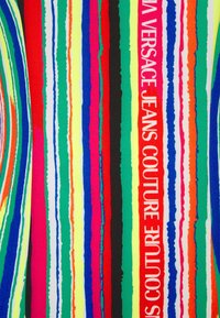 Bunt gemusteter Stoff mit vertikalen Streifen in verschiedenen Breiten und Farben, darunter Rot, Blau, Grün, Gelb und Schwarz. "VERSACE JEANS COUTURE" deutlich sichtbar.