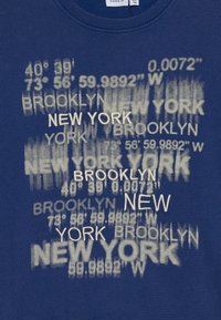 Blå hettegenser med hvit trykt tekst som viser geografiske koordinater og bynavnene "Brooklyn" og "New York" i varierende skriftstørrelser.