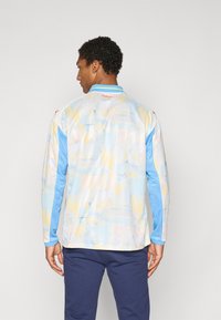 La camicia a maniche lunghe presenta un motivo astratto multicolore in tonalità pastello, con un colletto blu e maniche blu in contrasto. Dettaglio del logo rosso visibile.