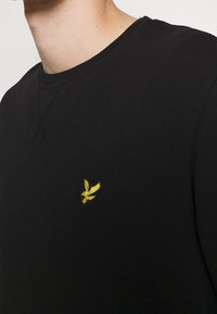 Svart långärmad sweatshirt med ribbad rund halsringning. Har en liten broderad gul fågel-logotyp på bröstet. Mjuk textur.