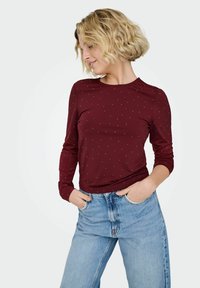 Kobieta o krótkich, kręconych blond włosach, ubrana w bordową bluzkę z długim rękawem w drobne kropki oraz jasnoniebieskie, wysokowodne dżinsy, z rękami w kieszeniach.