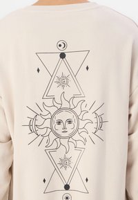 Maglione color crema con un design grafico nero raffigurante un sole e elementi celesti tra cui triangoli, diamanti e stelle.