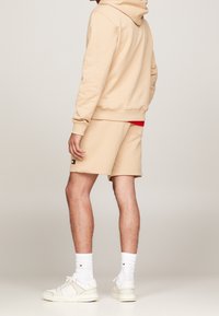 Beige hoodie en shorts van zachte stof, met een rode binnenlaag en logo-detail op de shorts. Gecombineerd met witte sneakers en sokken.