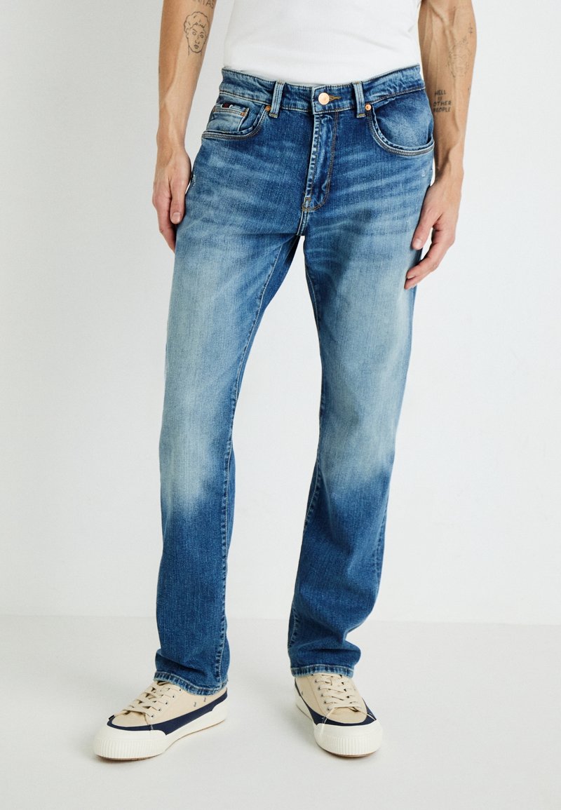 Homme portant un jean bleu délavé droit et des baskets blanches avec des détails bleu marine, debout devant un fond clair uni.