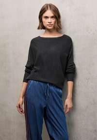 Maglione nero con vestibilità comoda e scollatura ampia, abbinato a joggers in denim blu con strisce laterali bianche e bordeaux.