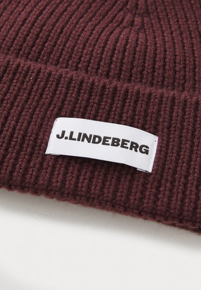 Burgundy ribbed knit beanie with a white fabric tag featuring "J.LINDEBERG" in black lettering, showing a textured finish.