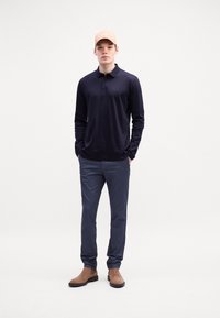 Marineblaue Langarm-Polohemd, geknöpfter Kragen, sandfarbene Baseballmütze, graue Hose und braune Wildleder-Stiefeletten. Einfaches Design, keine auffälligen Muster.
