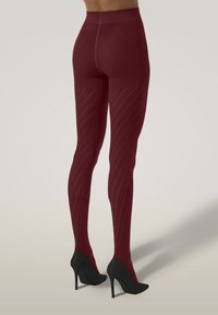 Person som bär burgundy mönstrade strumpor och svarta högklackade skor står mot en neutral bakgrund.