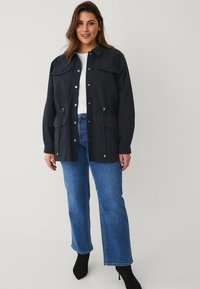 Giacca blu scuro a maniche lunghe con due tasche sul petto e vita regolabile, abbinata a jeans dritti blu chiaro e stivali neri alla caviglia.