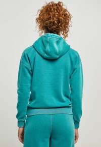 Tirkīza krāsas kapuce ar auklu, ribotām piedurknēm un apakšmalu. Izgatavota no mīksta materiāla ar brīvu piegriezumu, parādīta no aizmugures.