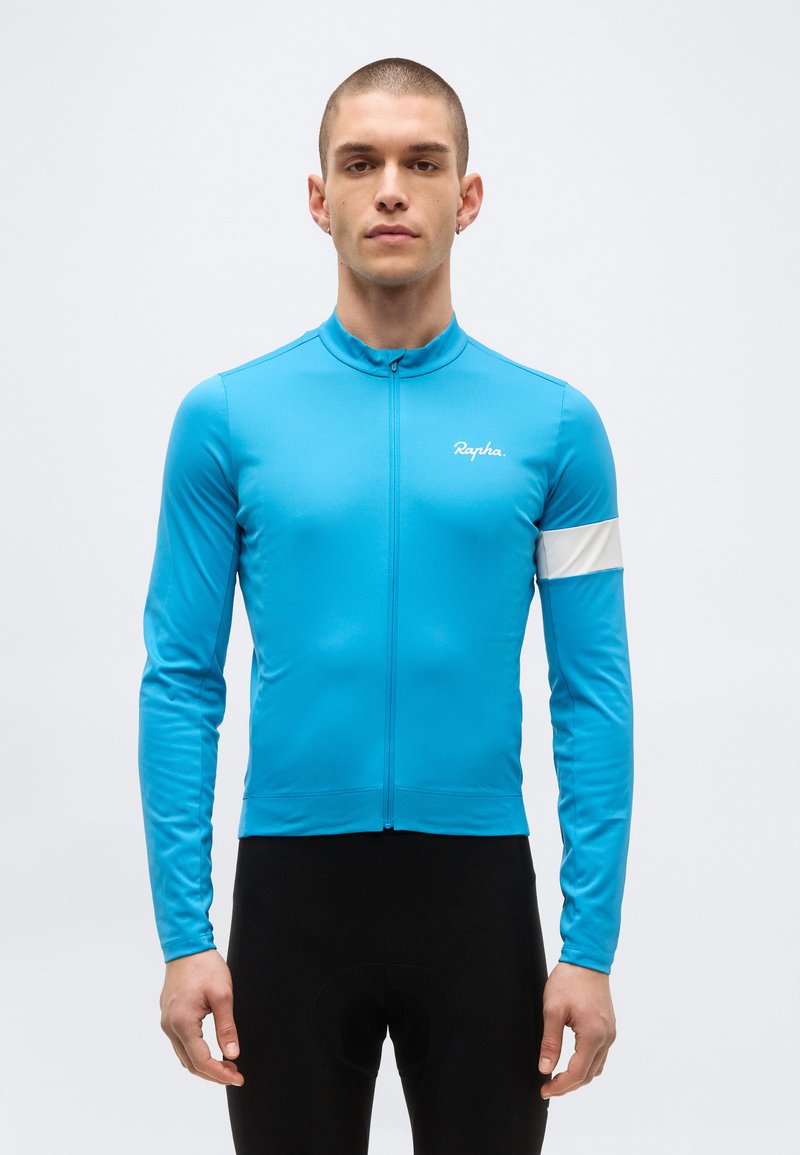 Νέος άνδρας φορόντας εφαρμοστό μπλε ποδηλατικό μπλουζάκι με λευκή ταινία στο μπράτσο και μαύρο ποδηλατικό σορτς, σε απλό φόντο.