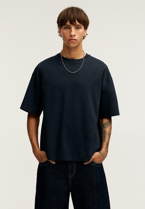 Jeune homme avec une coupe de cheveux brune courte portant un T-shirt noir oversize, un jean foncé, un collier en chaîne argentée et un bracelet, debout les mains dans les poches.