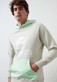 Sudadera gris claro con un cordón y un bolsillo en verde menta. Presenta una impresión gráfica circular con el texto "Monodock" en blanco. Textura suave.