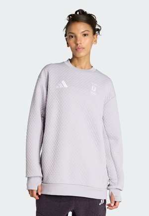 Frau trägt einen hellgrauen, strukturierten Pullover mit Adidas- und Olympiateam-Logos, kombiniert mit dunkler Hose, stehend vor einem einfarbigen Hintergrund.