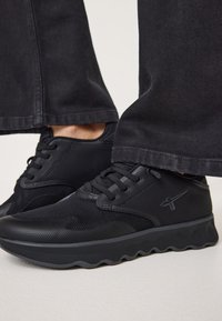 Czarne sportowe sneakersy z siateczkową cholewką, teksturowanymi akcentami i falistą podeszwą. Sznurkowe zapięcie i minimalne logo z boku.