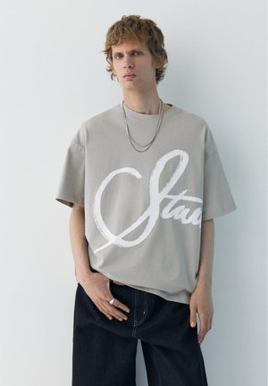 Młody mężczyzna noszący oversize beżowy t-shirt z dużym białym napisem, ciemne spodnie, srebrny łańcuszek z naszyjnikiem i pierścionek, stojący na tle jednolitej jasnej ściany.