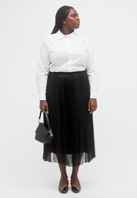 Γυναίκα με πλεξούδες στα μαλλιά, φορώντας άσπρο πουκάμισο με κουμπιά, μαύρη πλισέ φούστα, μαύρα επίπεδα παπούτσια και κρατώντας μια μαύρη τσάντα, κοιτάζοντας προς τα πλάγια.