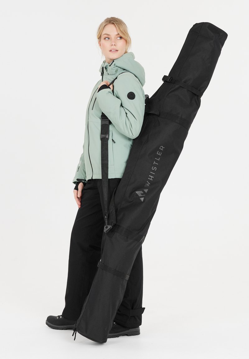 Mustat lumilautareppu, jossa on säädettävä hihna; tekstuuri kangas; näkyvästi esillä "WHISTLER" -logo; malli pukeutuneena mintunvihreään takkiin ja mustiin housuihin.