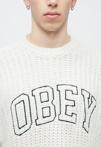 Pulover alb tricotat cu guler rotund costilat, având inscripția îndrăzneață neagră "OBEY" pe partea din față. Textură medie cu detalii de cusături lejere.