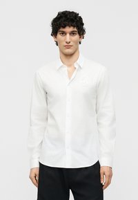 Νέος άνδρας με σγουρά μαλλιά, φορώντας λευκό πουκάμισο με κουμπιά και μικρό στρογγυλό λογότυπο στο στήθος, στέκεται μπροστά σε απλό φόντο.