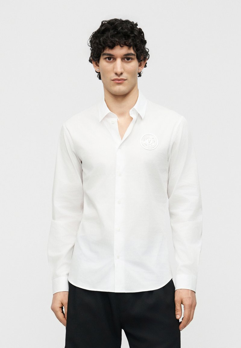 Νέος άνδρας με σγουρά μαλλιά, φορώντας λευκό πουκάμισο με κουμπιά και μικρό στρογγυλό λογότυπο στο στήθος, στέκεται μπροστά σε απλό φόντο.