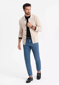 Hombre que lleva un cárdigan beige sobre una camisa negra, jeans azules, cinturón negro, reloj negro y zapatos negros, de pie y mirando hacia un lado.