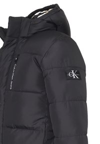Giacca a vento nera con cappuccio, dotata di una tasca con zip e una toppa con il logo sulla manica; texture liscia con sezioni trapuntate.