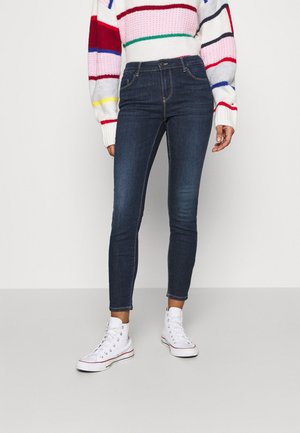 Mørkeblå skinny jeans med en glat tekstur, der har fem lommer og diskret syning. Sat sammen med hvide høj-tops sneakers.