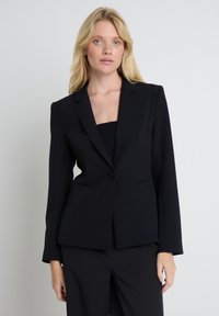 Szőke nő kék szemmel, fekete, testhezálló blézert és hozzá illő nadrágot viselve, egyszerű világos háttér előtt állva.