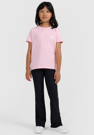 Chica con cabello negro que lleva una camiseta de manga corta rosa claro, pantalones negros y zapatillas blancas, de pie frente a un fondo gris claro.