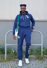 Sweatshirt azul de algodão com detalhes e logótipo amarelos, acompanhado de calças de fato combinando com texto. Ténis brancos e uma bolsa branca com alça verde.