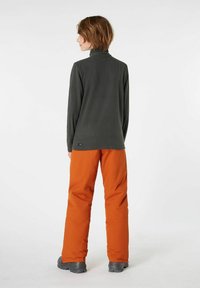 Haut long gris foncé à col montant, associé à un pantalon orange vif, vu de dos. Le matériau semble lisse avec une coupe décontractée.