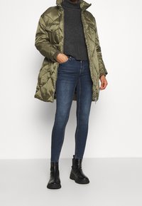 Abrigo verde oliva acolchado con patrón de diamantes, cuello alto, dos bolsillos en el pecho, llevado sobre un suéter gris oscuro y jeans ajustados azules, combinado con botas negras.