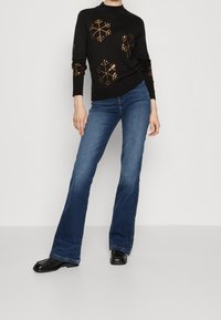 Pull en maille noire avec motif flocon de neige en feuille d'or, associé à un jean évasé bleu et des chaussures noires. Texture lisse et coupe décontractée.