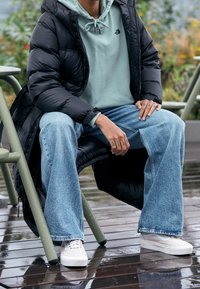 Giacca puffer nera sopra un hoodie verde chiaro. Jeans a zampa di elefante azzurri chiari e sneakers bianche. Seduto su un deck di legno con sedie verdi.