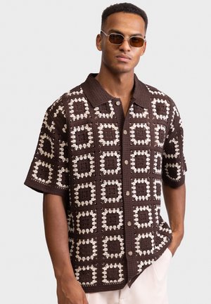 Uomo che indossa una camicia a maniche corte con bottoni, lavorata a uncinetto in marrone e crema, e occhiali da sole rettangolari con lenti marroni, in piedi con una mano in tasca.