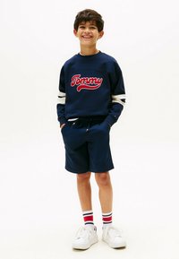 Lächelnder Junge mit den Händen in den Taschen, trägt einen marineblauen Pullover mit dem Aufdruck "Tommy", passende Shorts, gestreifte Socken und weiße Turnschuhe auf weißem Hintergrund.