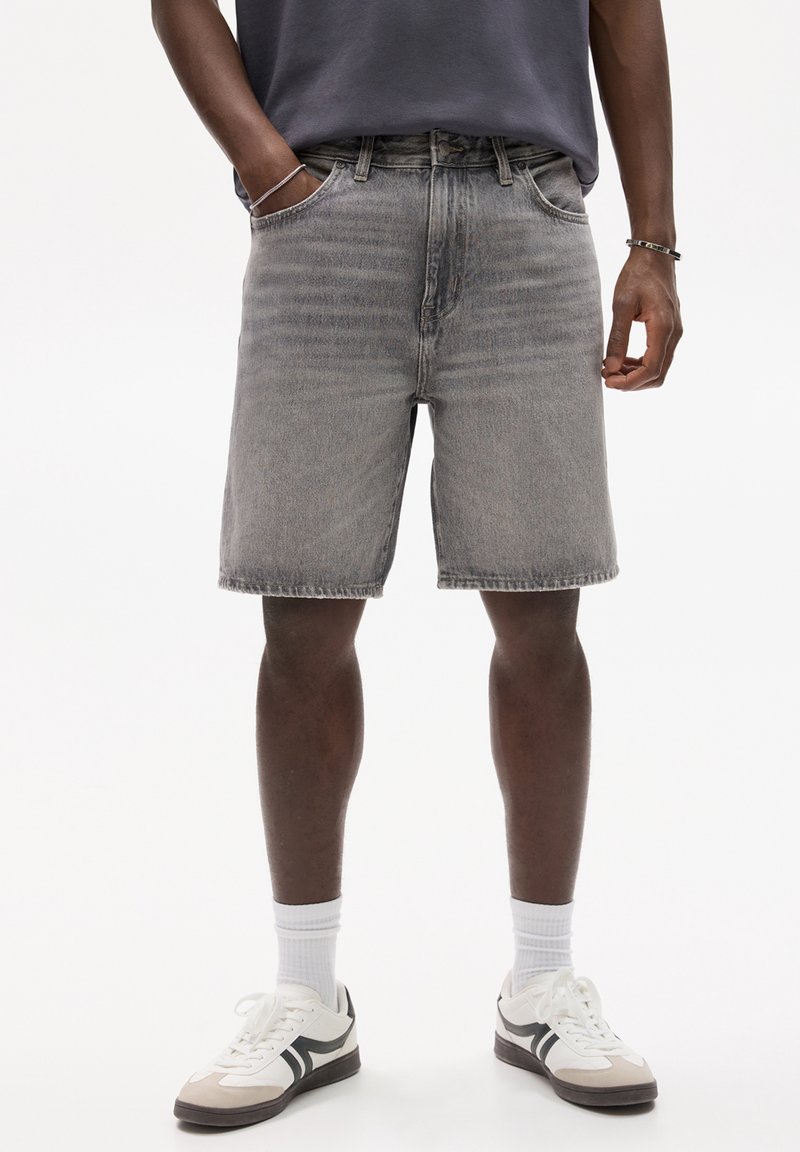 Personne portant un short en jean gris, des chaussettes blanches, des baskets blanches et noires, et un t-shirt gris foncé avec les mains le long du corps.