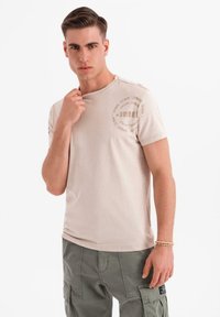Beige bavlněné tričko s kulatým výstřihem a nenápadným potiskem loga na rameni. Model má na sobě cargo kalhoty a stojí s jednou rukou na límci.