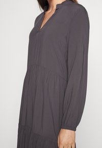 Femme portant une robe gris foncé à manches longues avec un tissu froncé à la taille et des poignets élastiques, debout devant un fond blanc uni.